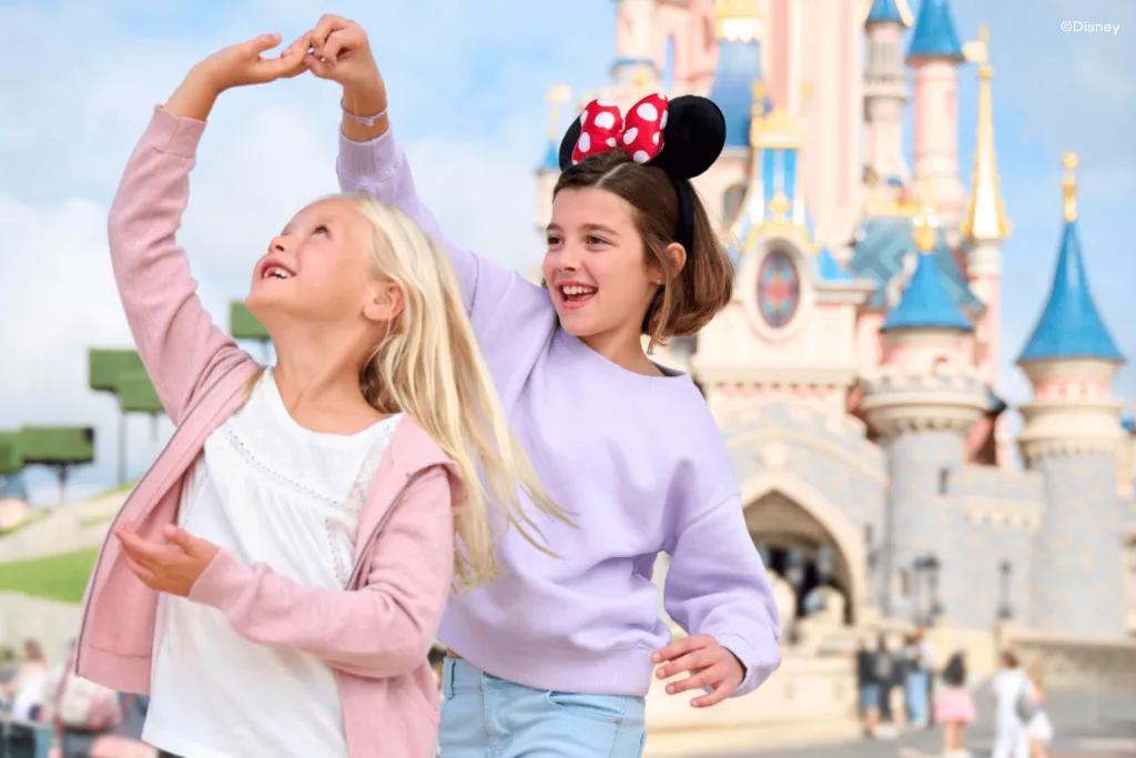 A girl holding hands above head, twirling another little girl around in front of a large blue and pink castle. There are guests stood behind walking towards the castle.