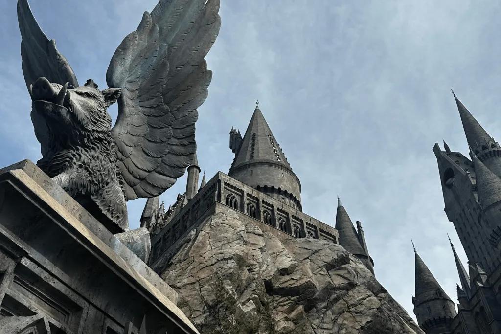Dark grey castle with pointed roofs sat on top of large rocks. To the left there is a statue of a pig with tusks and wings.