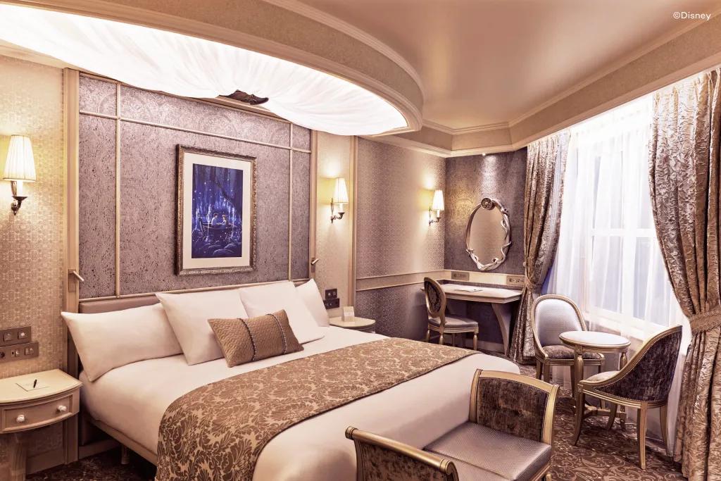 A bed with white and beige sheets and small cushions on. The room looks royal with a makeup table and a circular mirror above. There is a painting on the wall of Eric and Ariel in a boat holding hands.