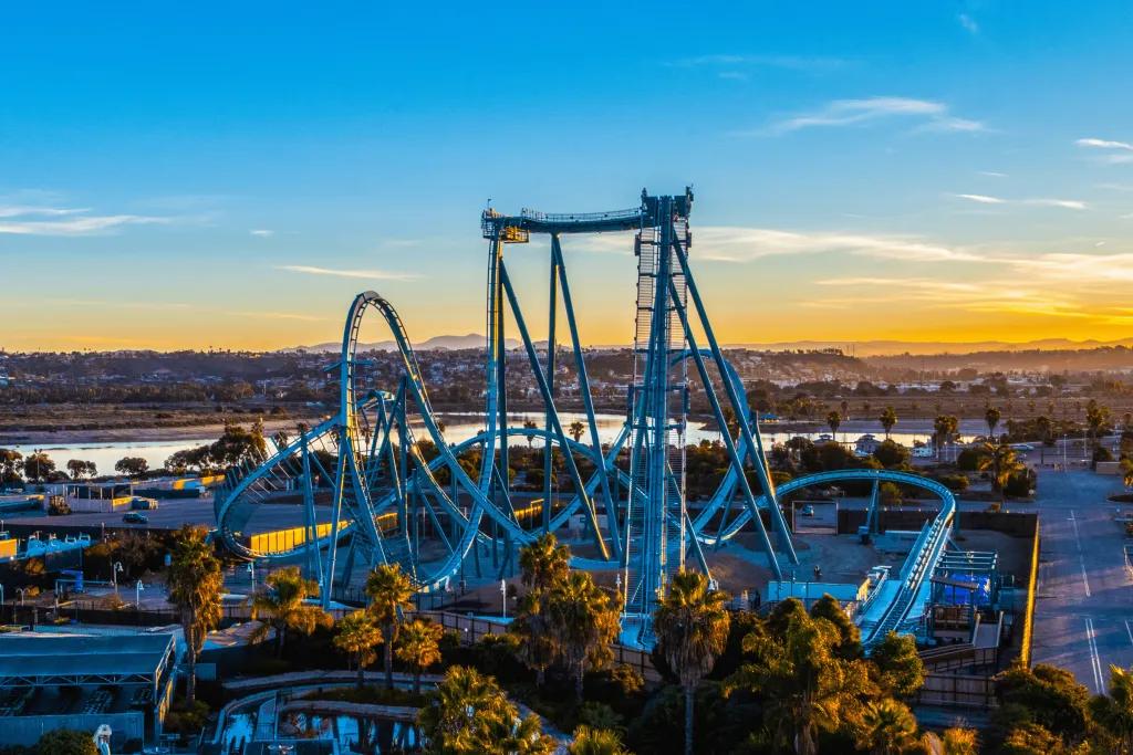Large light blue rollercoaster with steep drop and inversions. In the background there is a blue sky with a hint of orange sunset.