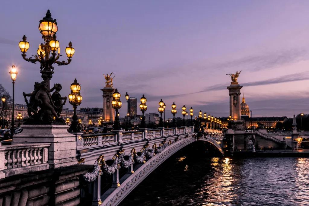 River with bridge over lined with black lamp posts lit up. There are magnificent artistic golden and stone statues.