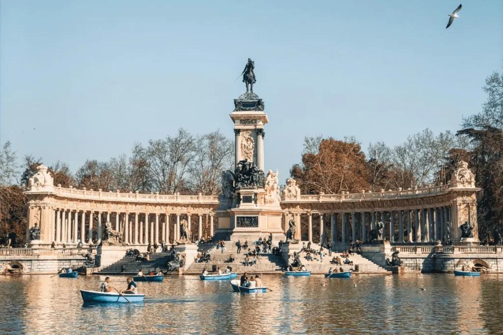Large stone pillars lined up behind a stone statue, on the top there us a man sat on a horse. There is a lake in front of it with couples sat in blue rowboats.