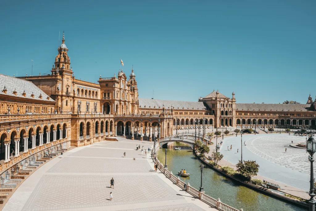 Grand historic building in Seville which is beige coloured with white detailing. There are many archways along the side held up by white pillars. There are spires, and there is a wide white walkway, with a river running alongside it, with white bridges over the river.