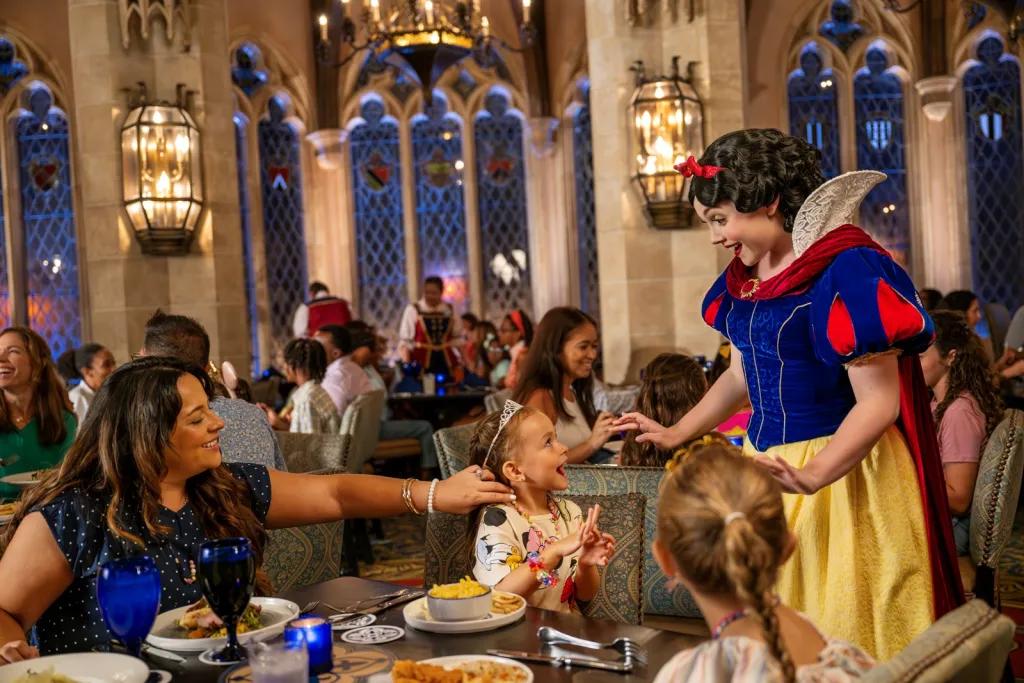 Mum and two girls sat a table eating. The mum is touching one of the girls hair as the little girl beams up at Princess Snow White who is at their table. Snow White is looking down smiling with a shocked expression. The small girl looks so surprised and happy. In the background there are tall windows on the brick walls and a large chandelier.