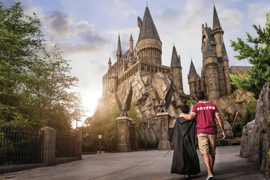A man wearing a red Griffindor shirt holding hands with a child in a black cloak and a wand. They are walking along an empty walkway in front of Hogwarts Castle.