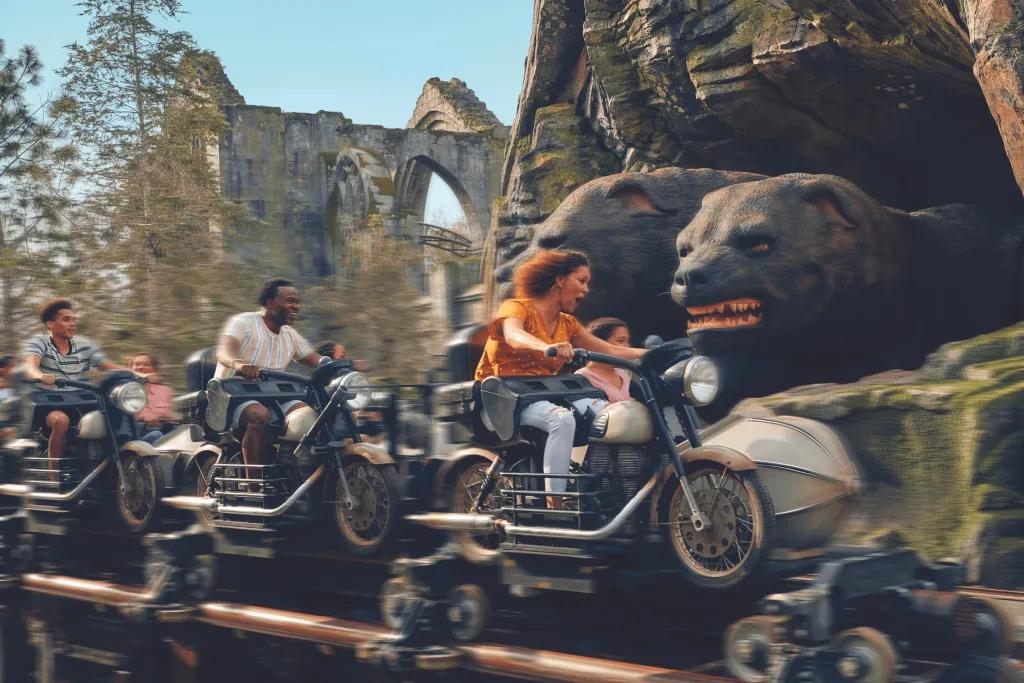 Guests sat on motorbike shaped rollercoaster vehicles in front of stone walls and a three headed dog sat on rocks.