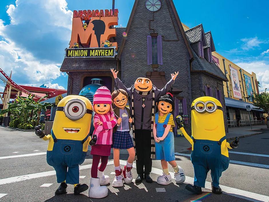 There is a black and grey house in the background with a large orange and yellow sign saying "Despicable Me Minion Mayhem" on. There are two minions in front with the Despicable Me characters (Gru, a man with a bald head, Agnes, a little girl with a black ponytail and dungarees on, Margo, a young brunette girl with glasses and Edith, a blonde child with a pink stripey hat on)