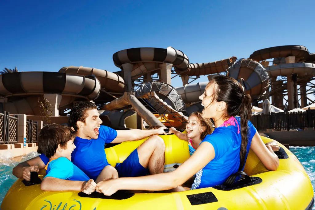 Group of four, 2 boys and 2 girls in a yellow circular rubber ring boat in a pool at the end of a beige and brown stripey waterslide.