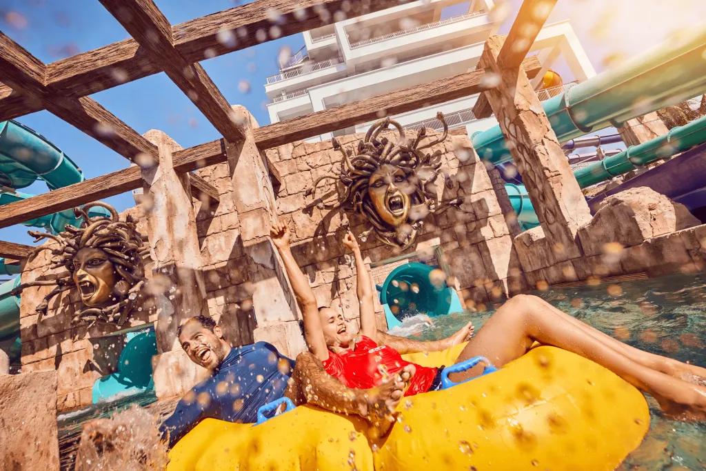 Man and woman in a double yellow rubber ring with water splashing around them. Behind them there are two waterslide exits coming out of a stone wall. Above there are golden face statues of Medusa.