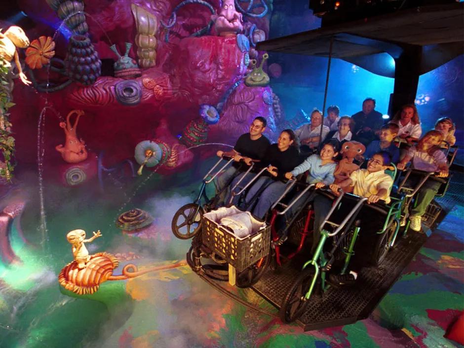 A hanging ride vehicle with bicycle shaped seating. Guests are sat on these smiling whilst looking at the immaculately themed surroundings, yellow, pink and blue alien figures and flowers.