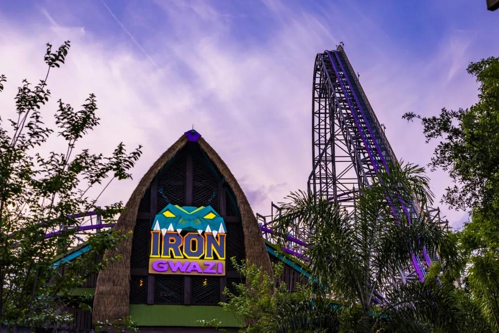 Iron Gwazi sign with green snake cartoon on top. In the background there is a tall purple roller coaster hidden behind trees and the sky is also a pink/purple shade.