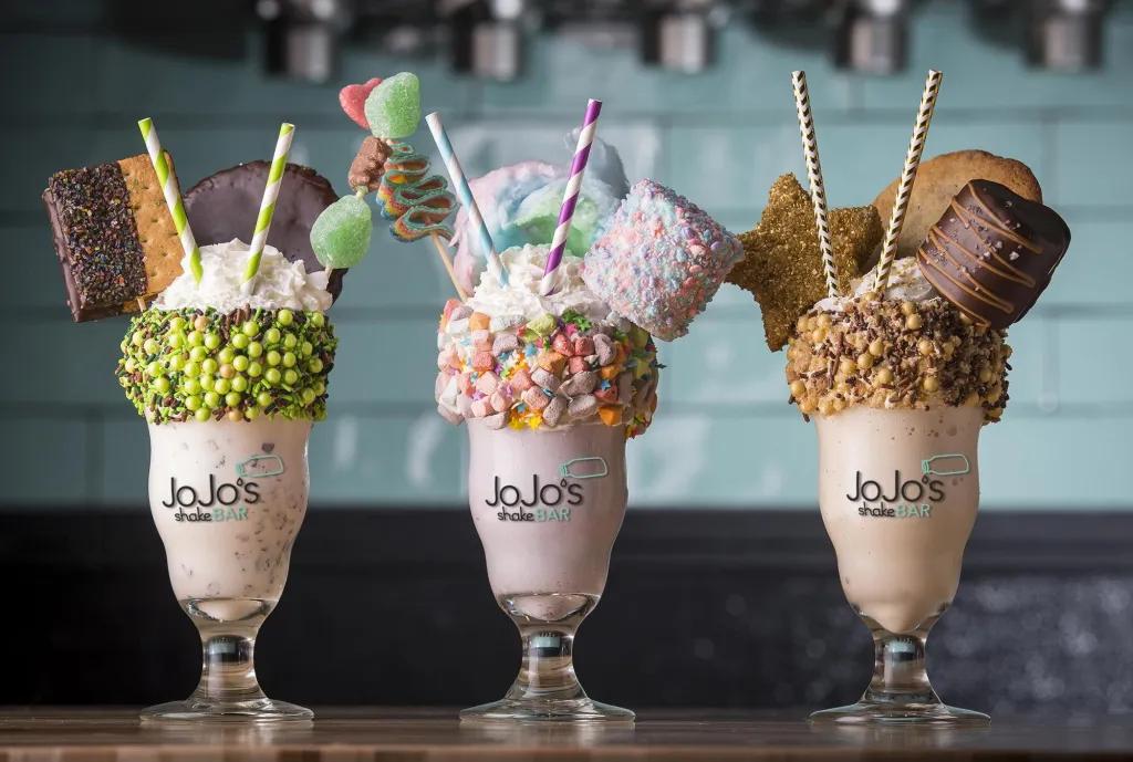 Three huge milkshakes in glasses lined up, one with green balls lining the top of the glass, a pink drink and two large biscuits sticking out with a cocktail stick of sweets and two green straws. The second one has pink and orange sweets lining the glass, a pink liquid with whipped cream, a giant marshmallow on a stick, pink and blue cotton candy and a stick with sweets on. Finally the last one has brown chocolate balls on the top of the glass, a chocolate flavoured milkshake and biscuits.