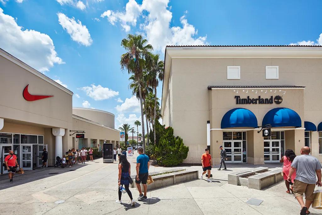 People walking through rows of shops on either side lined with palm trees. People are holding shopping bags and there are shops such as Timberland and Nike.