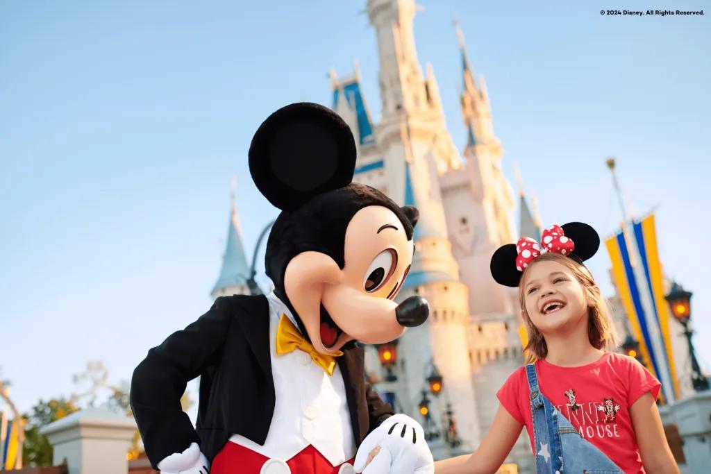 Mickey Mouse, a cartoon mouse wearing a yellow bow tie and a suit coloured red and black, holding hands with a young girl wearing Minnie Mouse ears stood in front of a white and blue castle at sunrise.