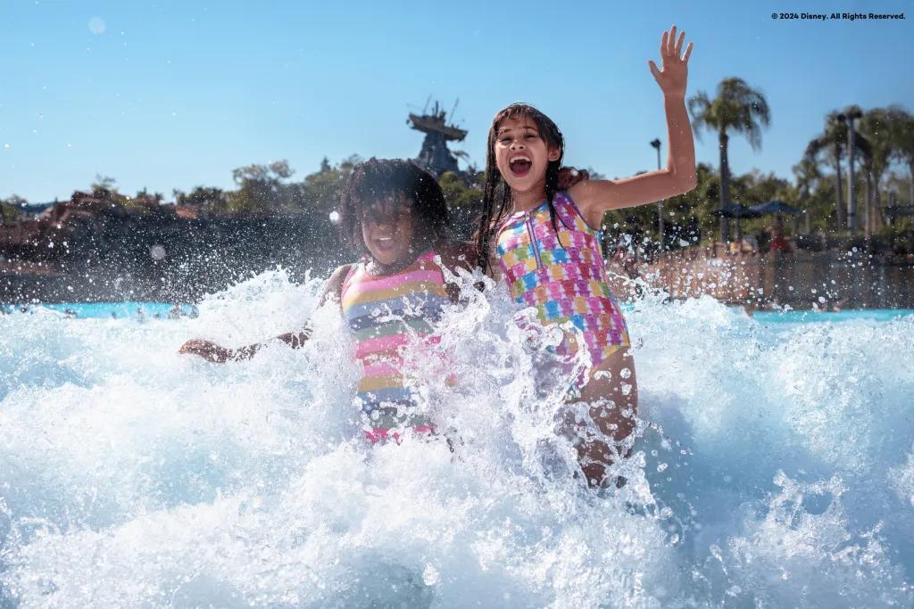 Two children smiling and laughing as they run out of a large pool with their hands in the air. There is water splashing around them and there is a statue of a boat stranded on top of a large rock in the background.