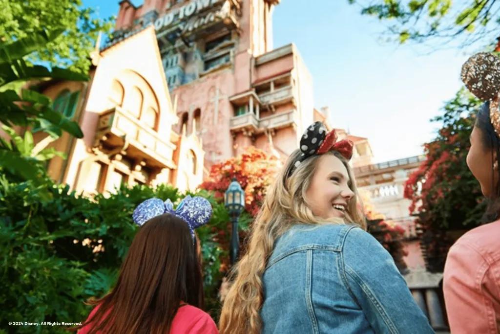 A group of young girls stood wearing mickey ears underneath a large dishevelled looking tower which is the Tower of Terror. One of the girls is turned to the others smiling. 