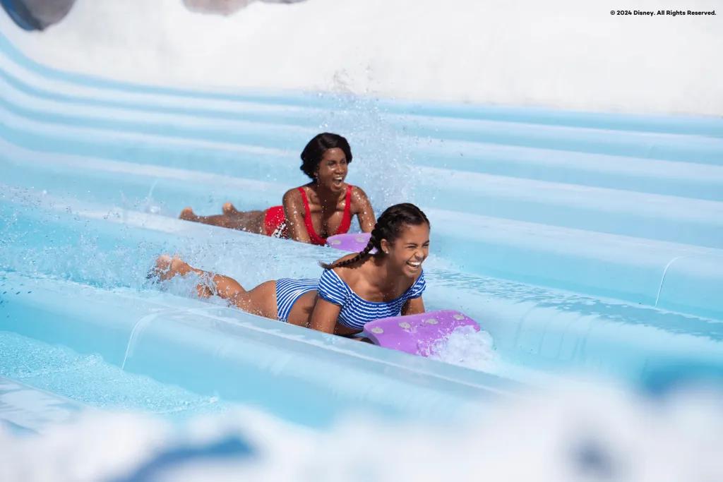 There is a mum and daughter sliding on mats down a light blue waterslide side by side. There are seven in a row for people to race down. Both are smiling.