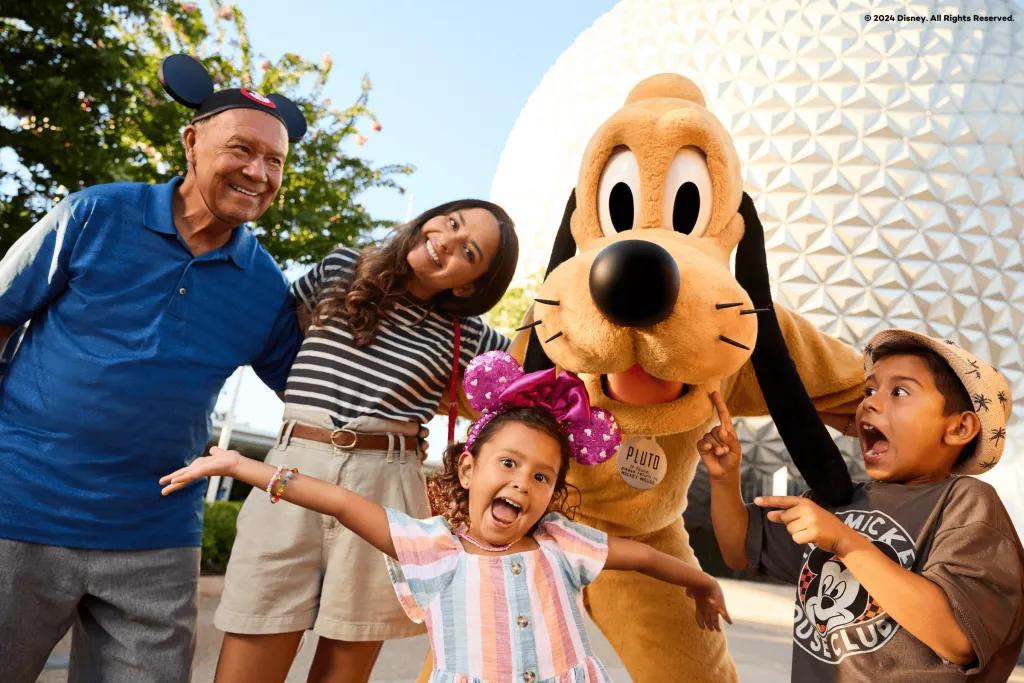 There is a golf ball white shaped building in the background. There is a yellow cartoon dog called pluto stood with a family of four. The young boy is gasping pointing at his sister in shock, the daughter is stood in front with pink sparkly ears with her arms extended out, and the parents are stood arm in arm smiling.