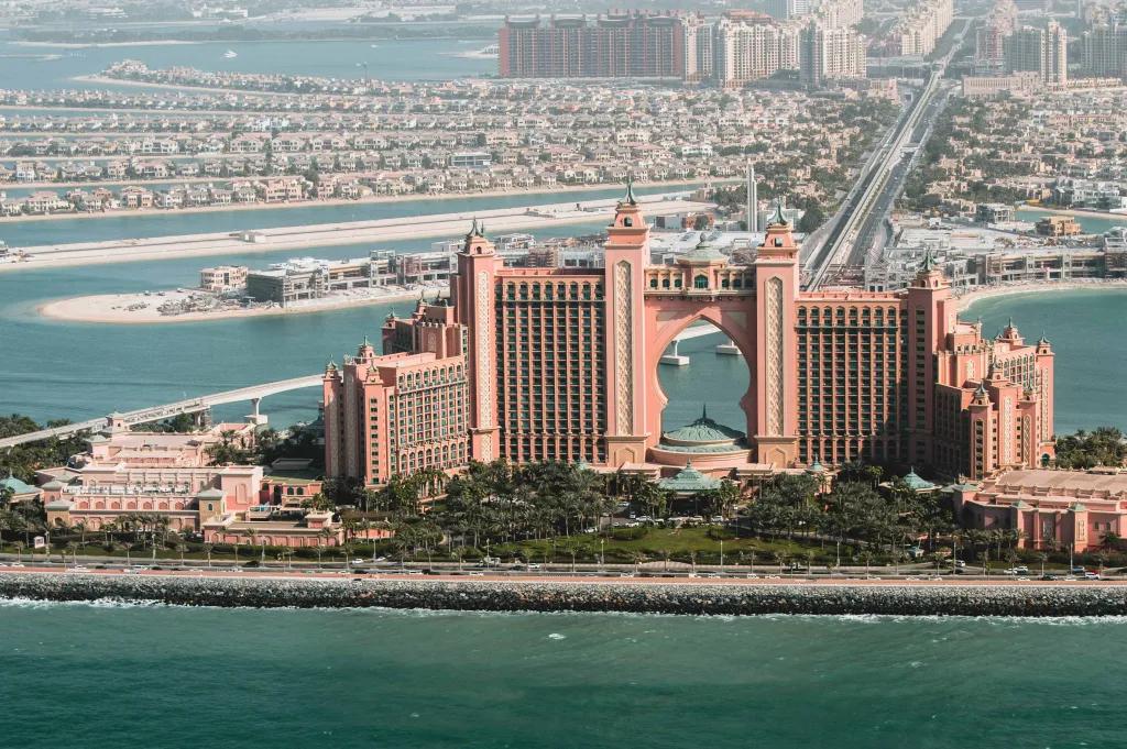Coral pink large hotel staggered levels and in the middle is a huge arch cut from the building. There is water surrounding this hotel and a long road connecting it to mainland Dubai.
