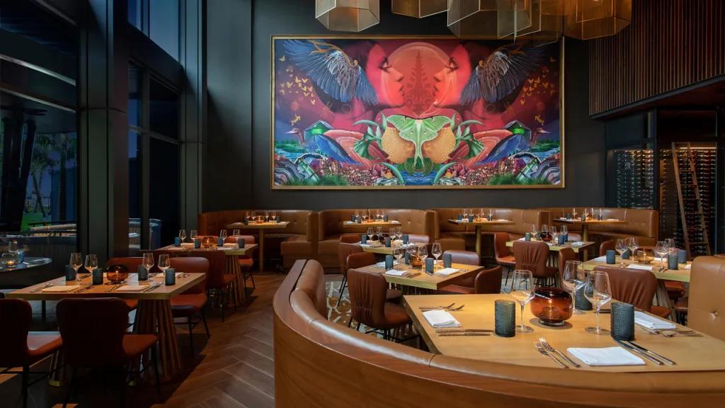 Dimly lit restaurant with round booths and large tables. There is a bright piece of artwork on the wall which is bright red, blue and purple which stands out amongst the dark wooden 