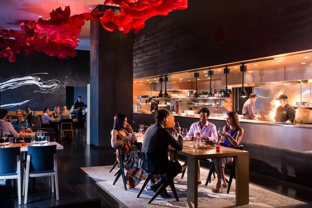 Modern restaurant with four people sat around a table talking. Above there is an artistic reg light shaped like a cloud. Behind there is a kitchen with chefs and flames coming from frying pans.