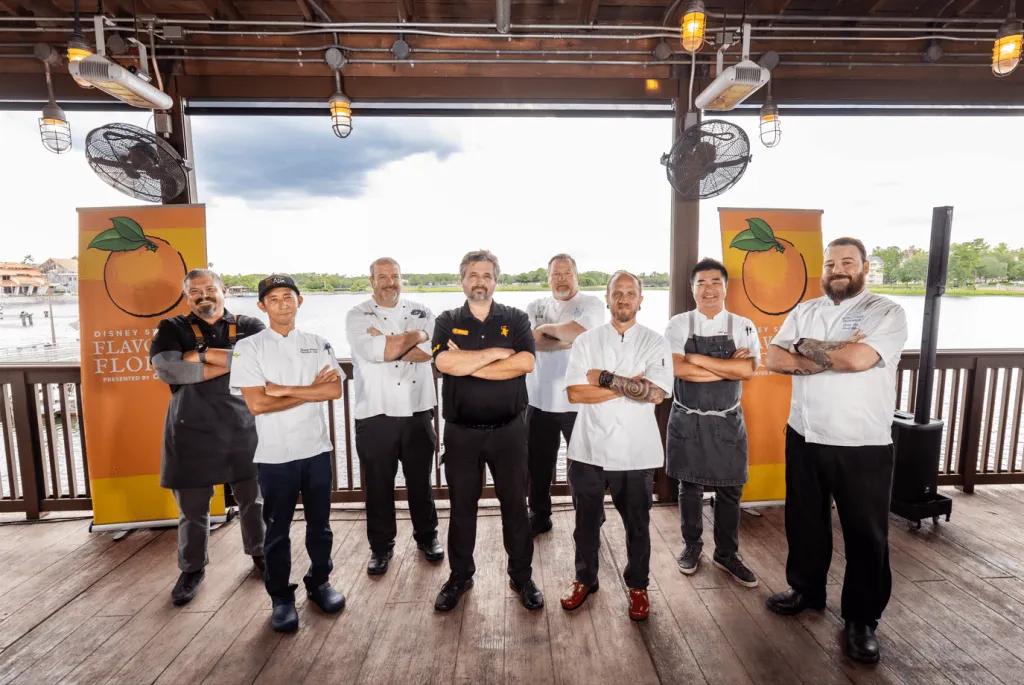 9 chefs standing on a wooden stage facing the camera with their arms crossed