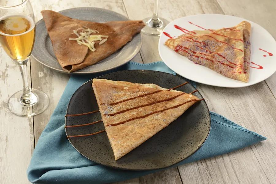 Three plates of crepes and a glass of champagne on the side. One crepe has chocolate sauce on it, one has strawberry sauce and the other has white chocolate shavings.