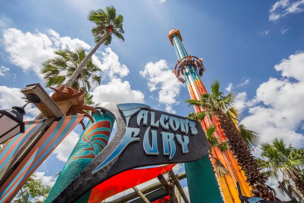 A sign which says Falcon's Fury and there is a ride in the background which is a large drop tower with the ride in motion and the circular seating dropping through the sky.