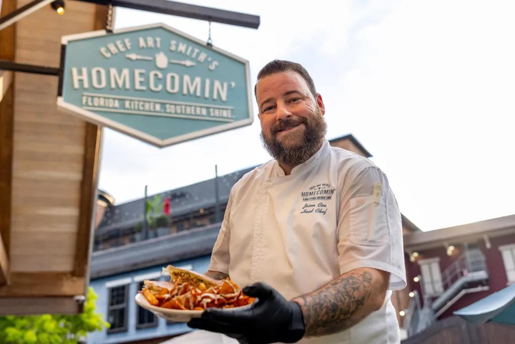 A chef holding a plate of food and smiling at the camera