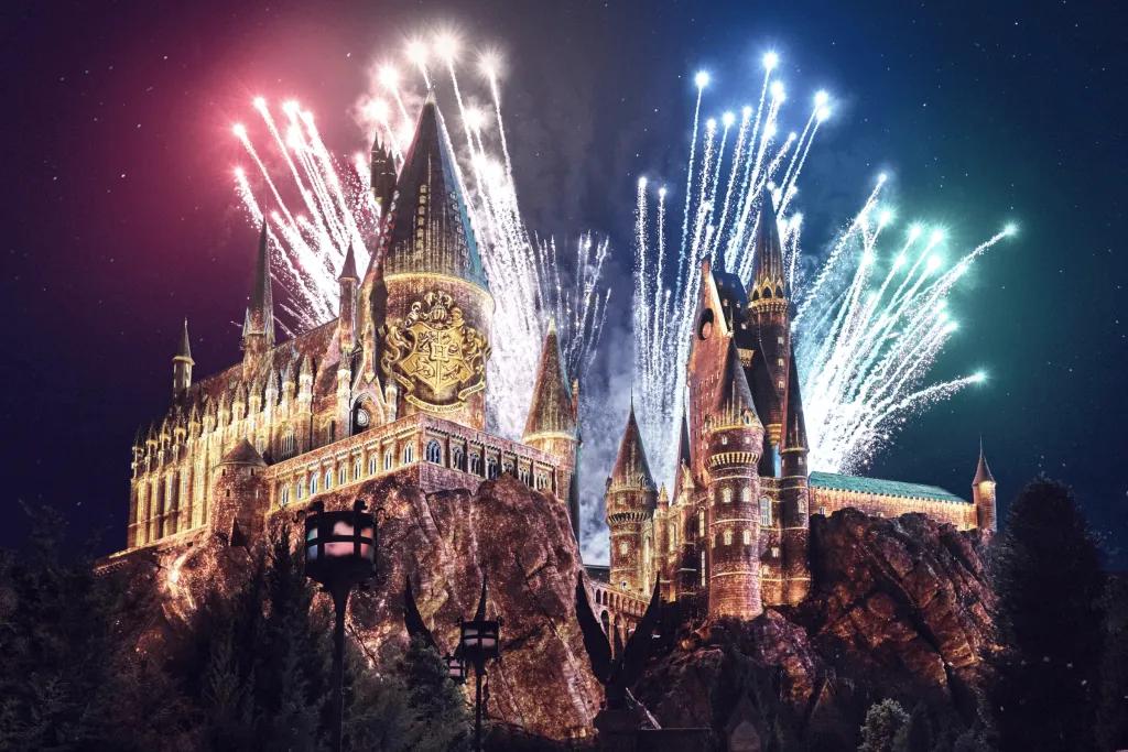 Large brown castle sat on top of rocks with many towers. It is night time and there are fireworks shooting out behind the castle of different colours, red, yellow, blue and green to match the house colours. There are also projections on the castle with it lit up yellow and a large shield with four different animals on it.