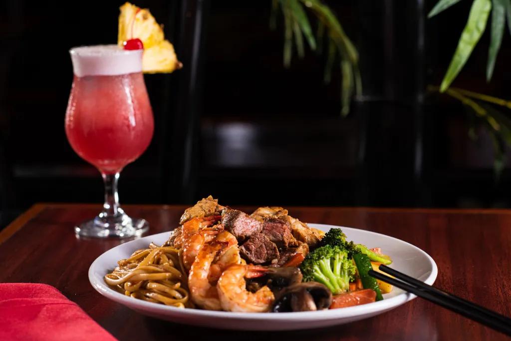 White plate with steak, prawns, noodles and broccoli on it mixed together. There is also a cocktail full of red liquid and a slice of pineapple on the glass.