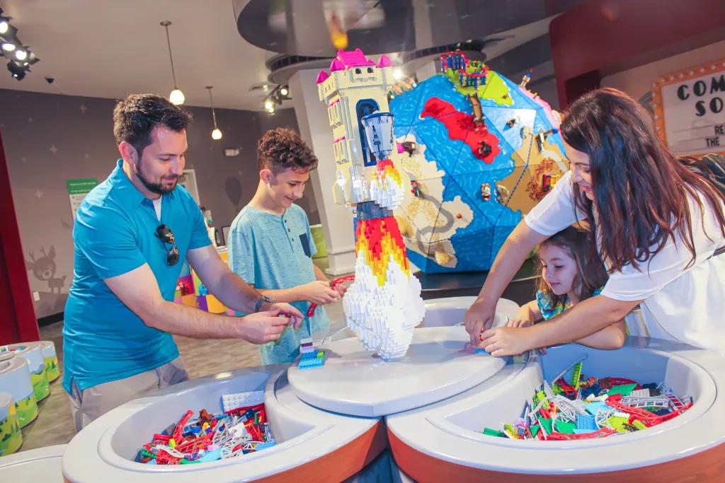Family of four crowded around a lego table building their own creations. The mum and daughter are stood together and the dad and son are stood on the opposite side. In the background there is a lego earth.