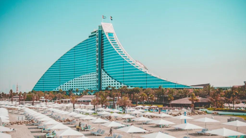 Building shaped like a sail made purely of windows. On top there are two UAE flags, below there is a beach with pristine white sand and white umberellas.