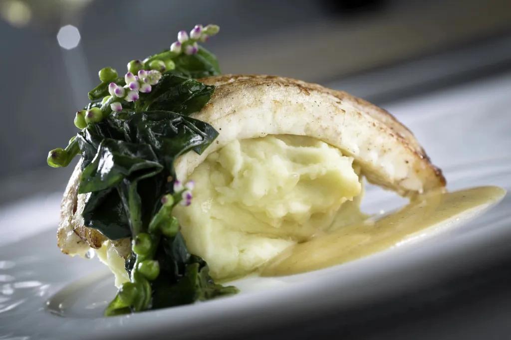 Slice of fish placed on mash potato scoop. There's green vegetables on top and a beige coloured gravy.