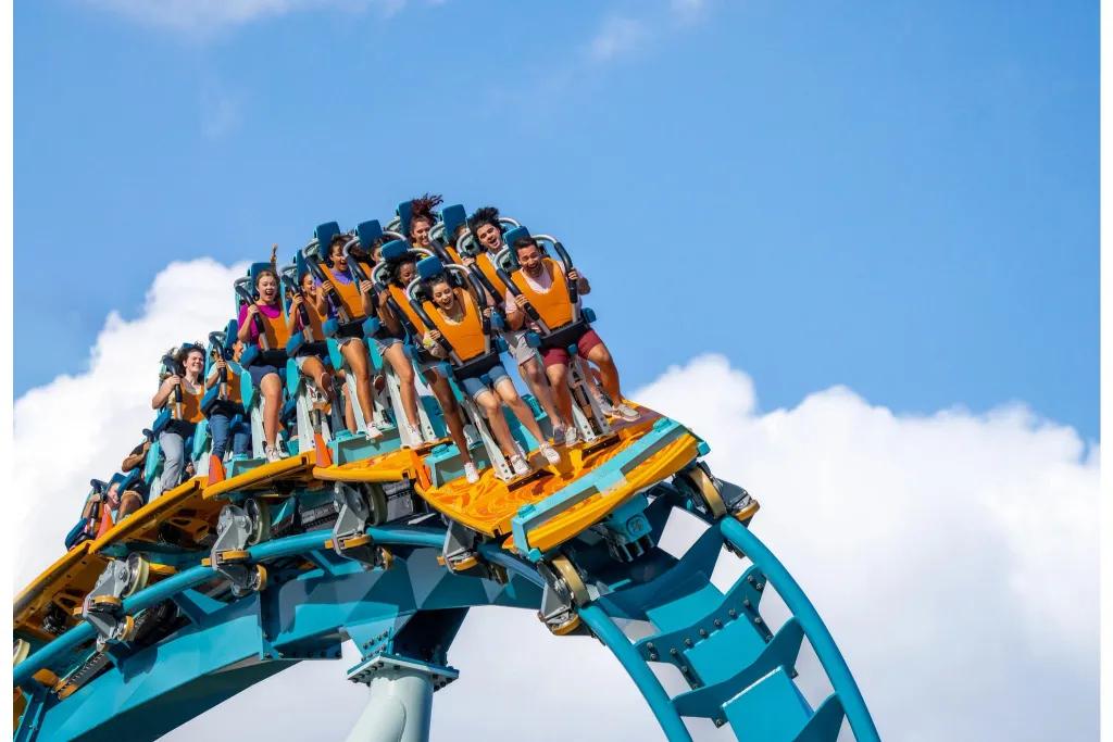 People on a rollercoaster car standing up with harnesses around their shoulders. They are stood on a vehicle which is themed as a surfboard coloured orange and turquoise with the track a light blue.