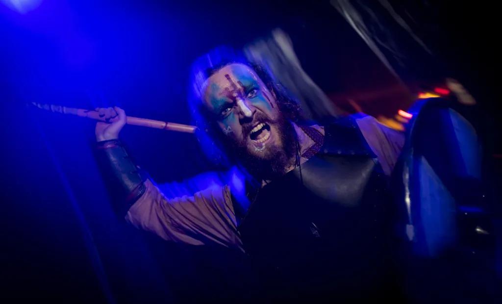 A dark and blurry image of a man screaming whilst holding an axe which he looks like he's about to hit the camera with. He has face paint on and an apron. 