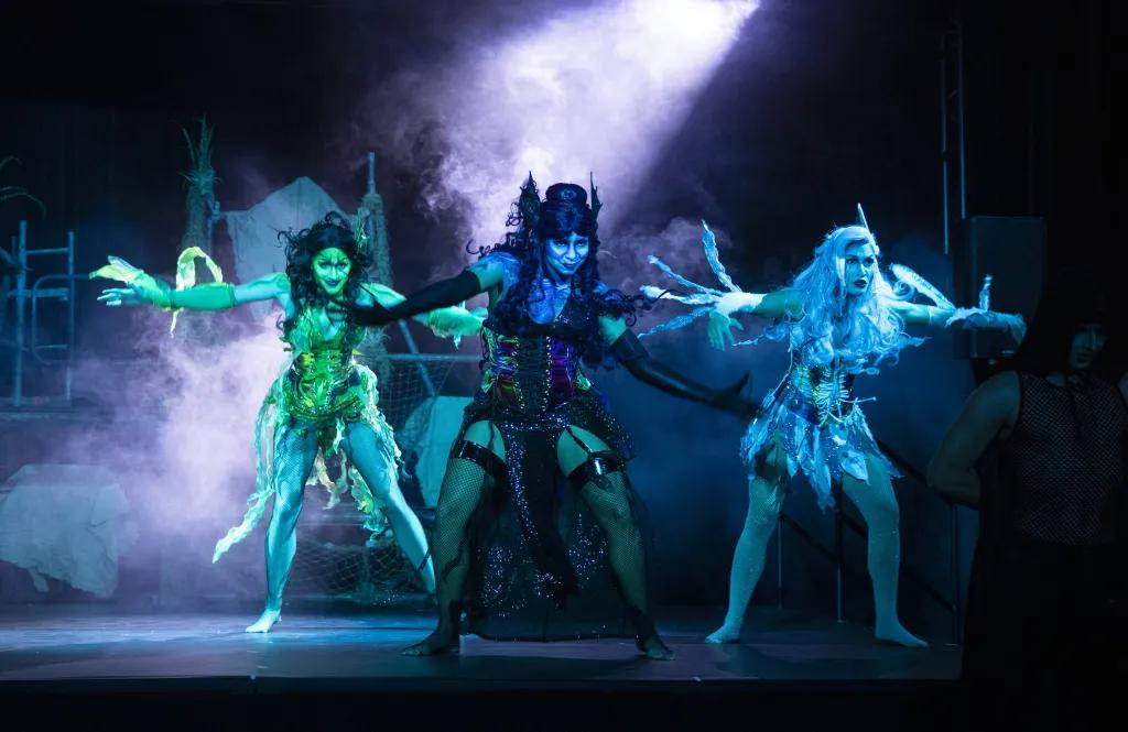 Three women dressed as sirens in blue lighting, one green, one black and blue and the other icy blue. They are wearing textured clothes and have matching hair. Each of them are in the middle of a dance with their arms out reached.