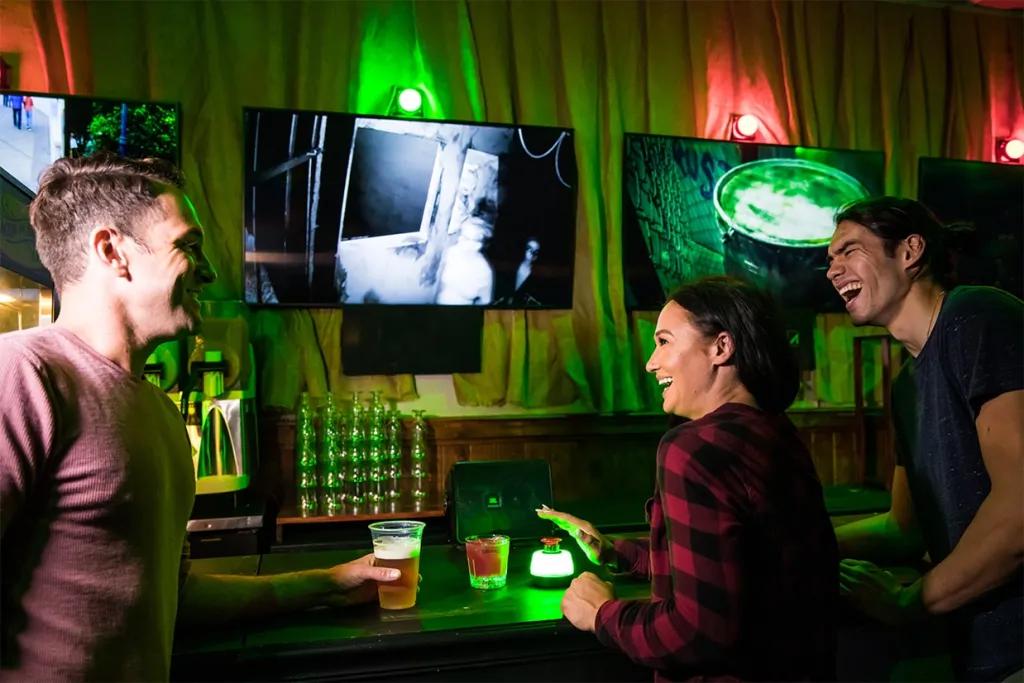 Room with curtains and green and red lighting above TV's which look like they are playing CCTV footage. There are three adults sat with drinks all laughing with one woman holding her hand above a red button with white lighting around it.
