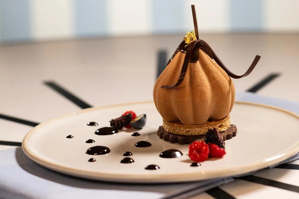 Plate with mousse shaped like a tulip on a biscuit and chocolate shavings on. There are also drops of chocolate sauce around the plate.