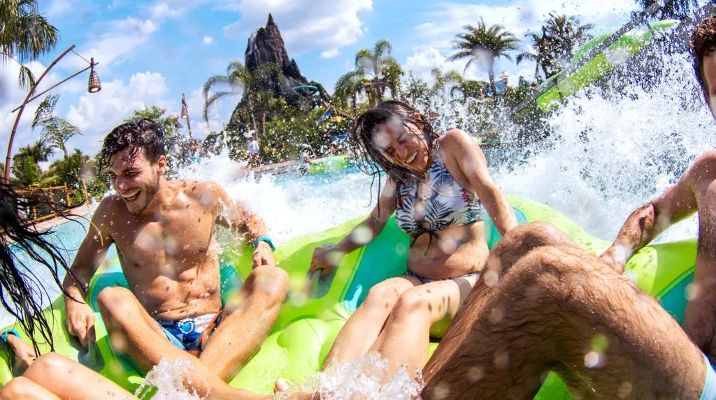 Four young adults sat on a green rubber ring splashing into a large pool with a large statue of a volcano in the background.