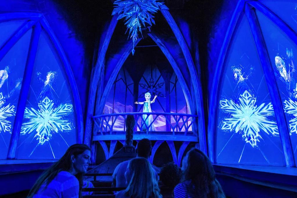 Queen Elsa stood on ice balcony surrounded by ice walls and projections of snowflakes. There is a chandelier above hanging made of ice. Below there are guests sat in rows on boat.