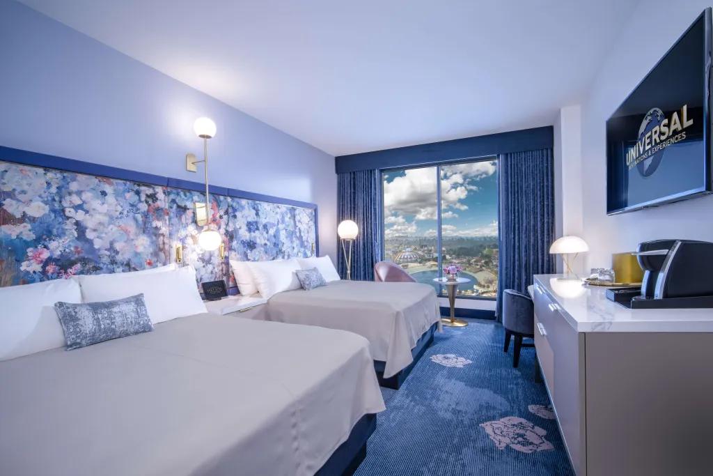 Blue room with two double white beds side by side in spacious area. There is a tv on the wall with the Universal sign on and there is a big window with view of Epic Universe. On the wall there is a floral pattern in a blue hue.