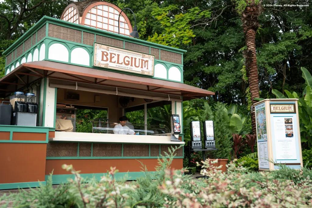 A food stand designed in white and green colours. There is a sign on top saying Belgium in orange. Inside there is a man in a white uniform.