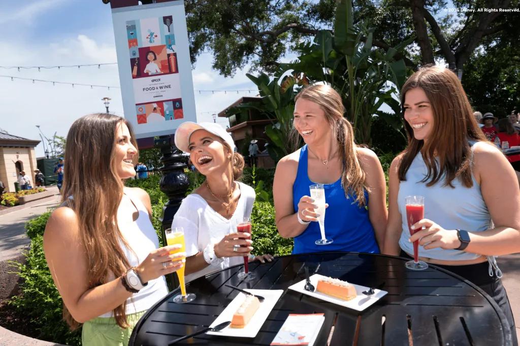 Four girls looking at one another laughing as they are sat a table holding different coloured plastic glasses of drinks. Two are red, one yellow and one white. There are plates of perfect looking desserts on the table with plastic knives and forks.