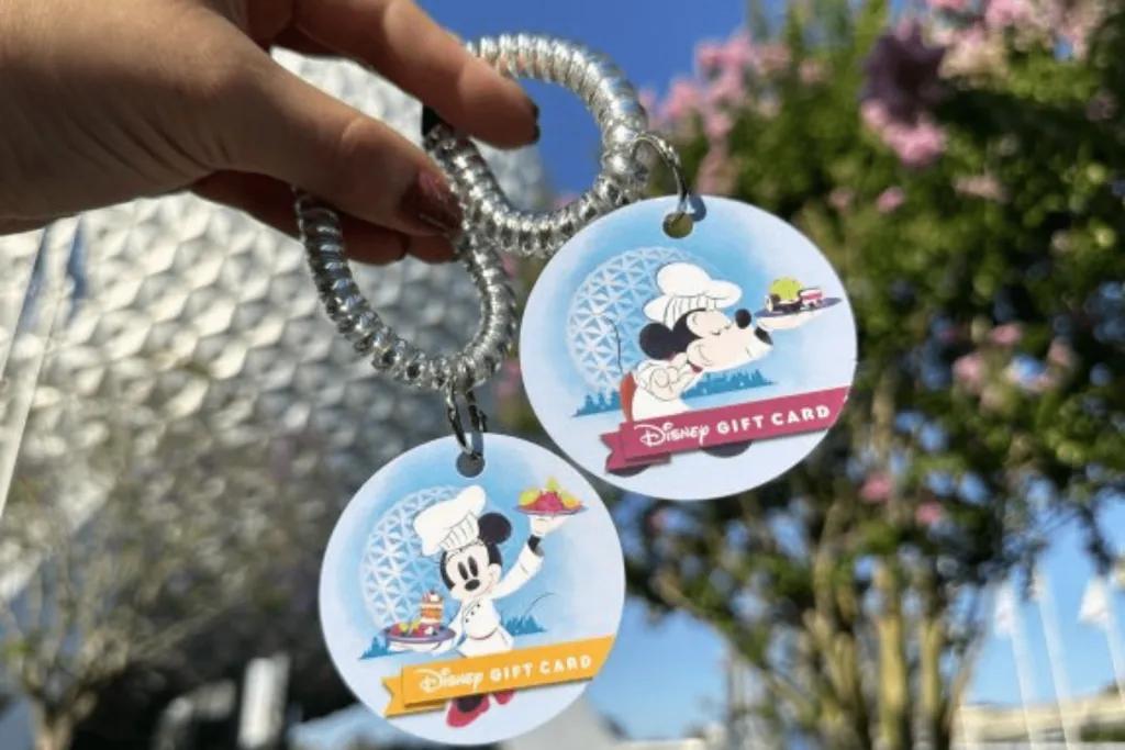 Two see through wristbands held up by a hand, on each are designs of blue backgrounds and one has Mickey Mouse and the other has Minnie Mouse. Both characters are wearing chef costumes and holding meals. Text on it says Disney Gift Card. Spaceship Earth is in the background.