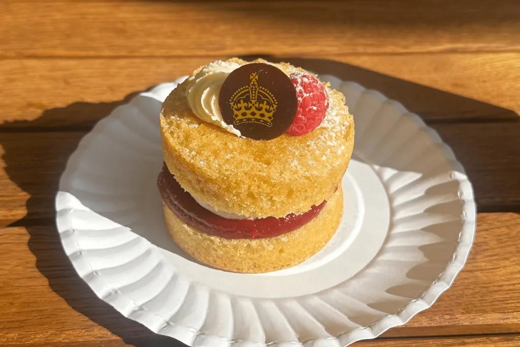 Small, circular Victoria sponge cake served on paper plate. It is two vanilla cake slices with cream and jam in the middle. On top there is cream, a raspberry and a circular piece of chocolate with golden image of crown on it.