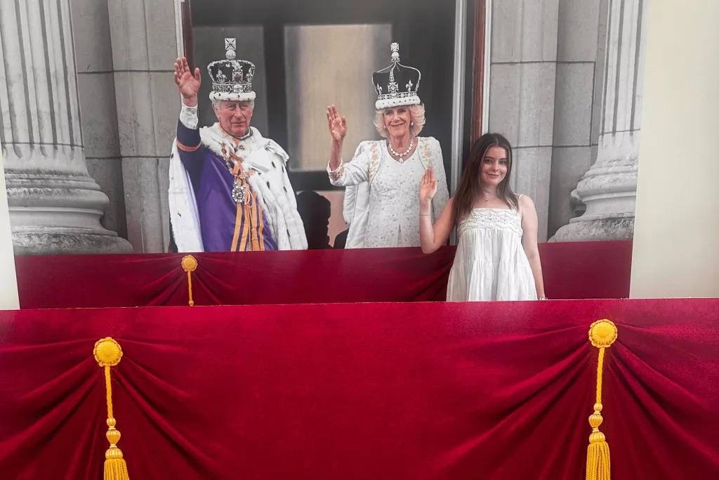 Girl stood in front of image of King Charles and Queen Camilla stood on the balcony during coronation with crowns on their heads. They are both waving and so is the girl waving and smiling. There is also a cardboard red velvet curtain in front to replicate balcony.
