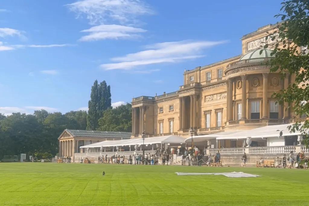 The back of Buckingham Palace, a large beige building which backs onto large green grass patch and trees. There is also a white tent with cafe in.