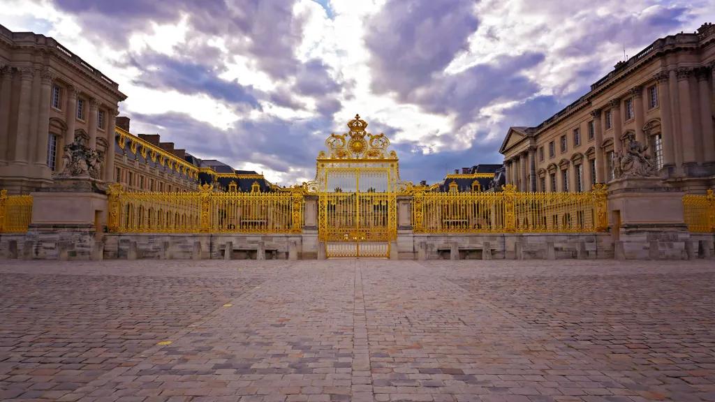 Golden gates with beautiful royal details, there are stone statues and behind is a grand palace.