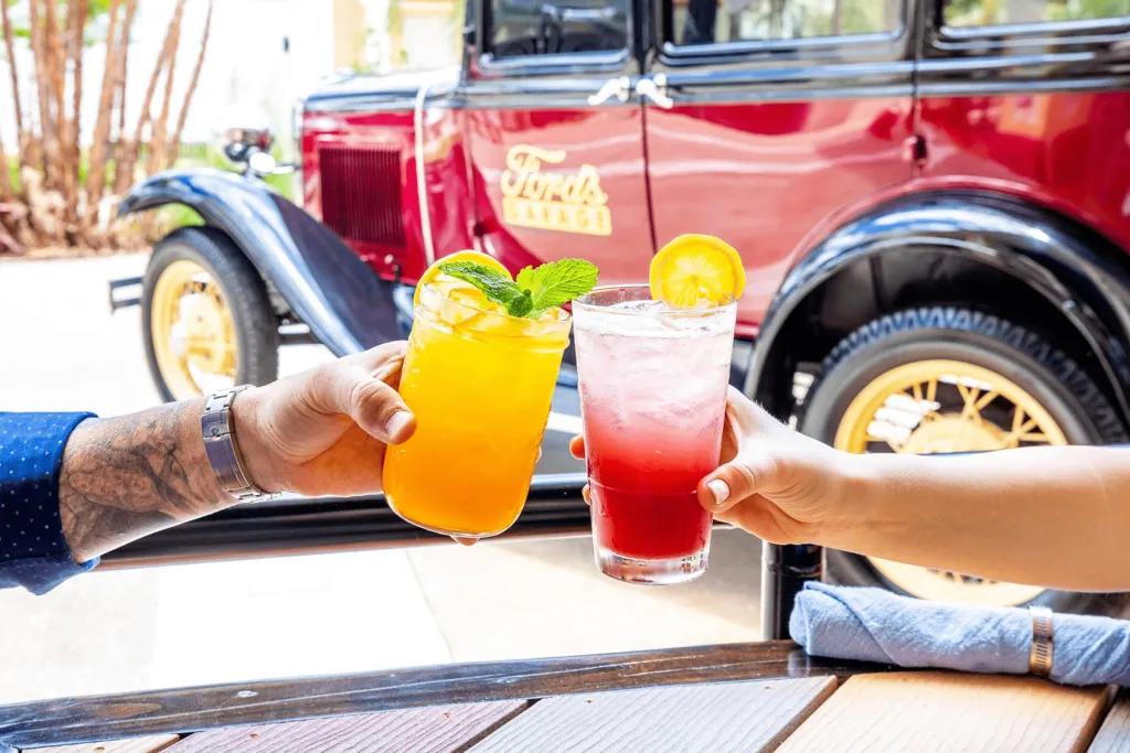 Two people clinking glasses of cocktails in front of an old fashioned car with Ford's Garage on the side